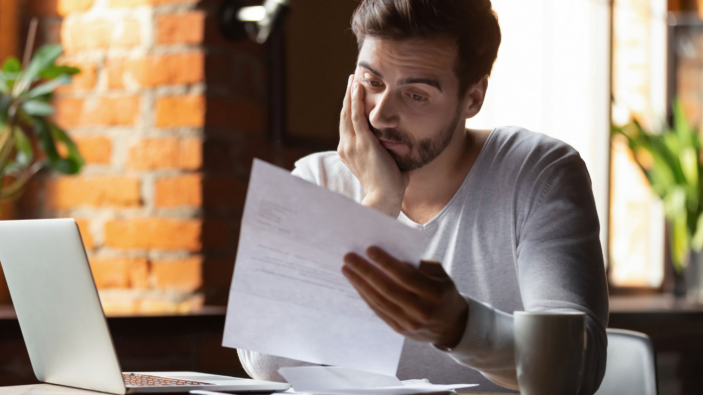 Young man sitting at laptop hold account statement with hand on face