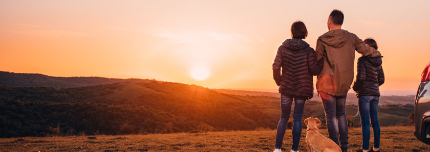Family on road trip standing by car watching sunset over hill