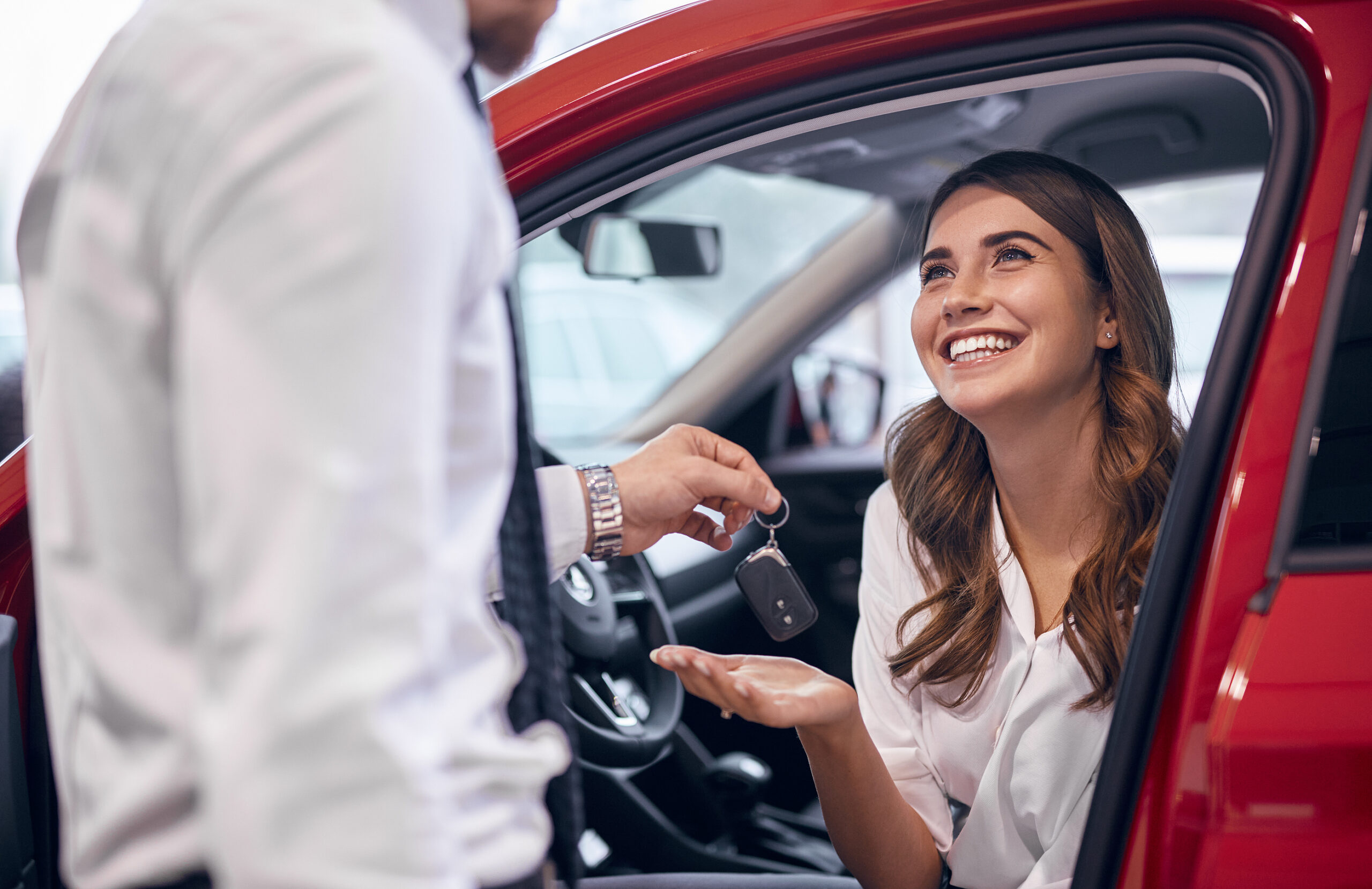 A smiling woman seated in a red car receives the keys to her new vehicle after determining the down payment on her car loan