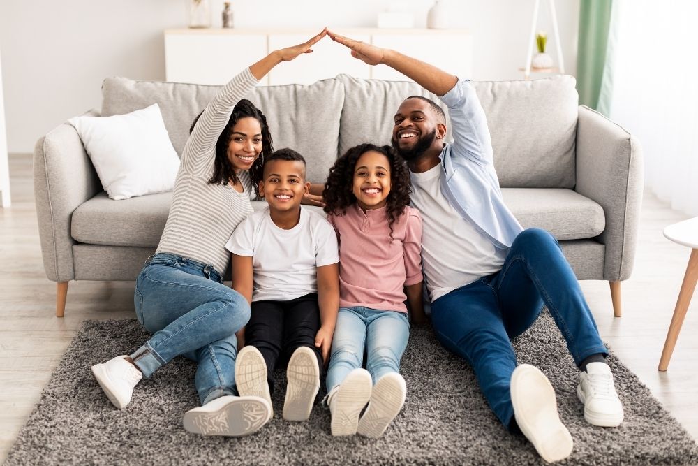 a family of four sits on the floor in front of their couch, the parents hold their hands up to make a roof over their children's heads, representing their home equity