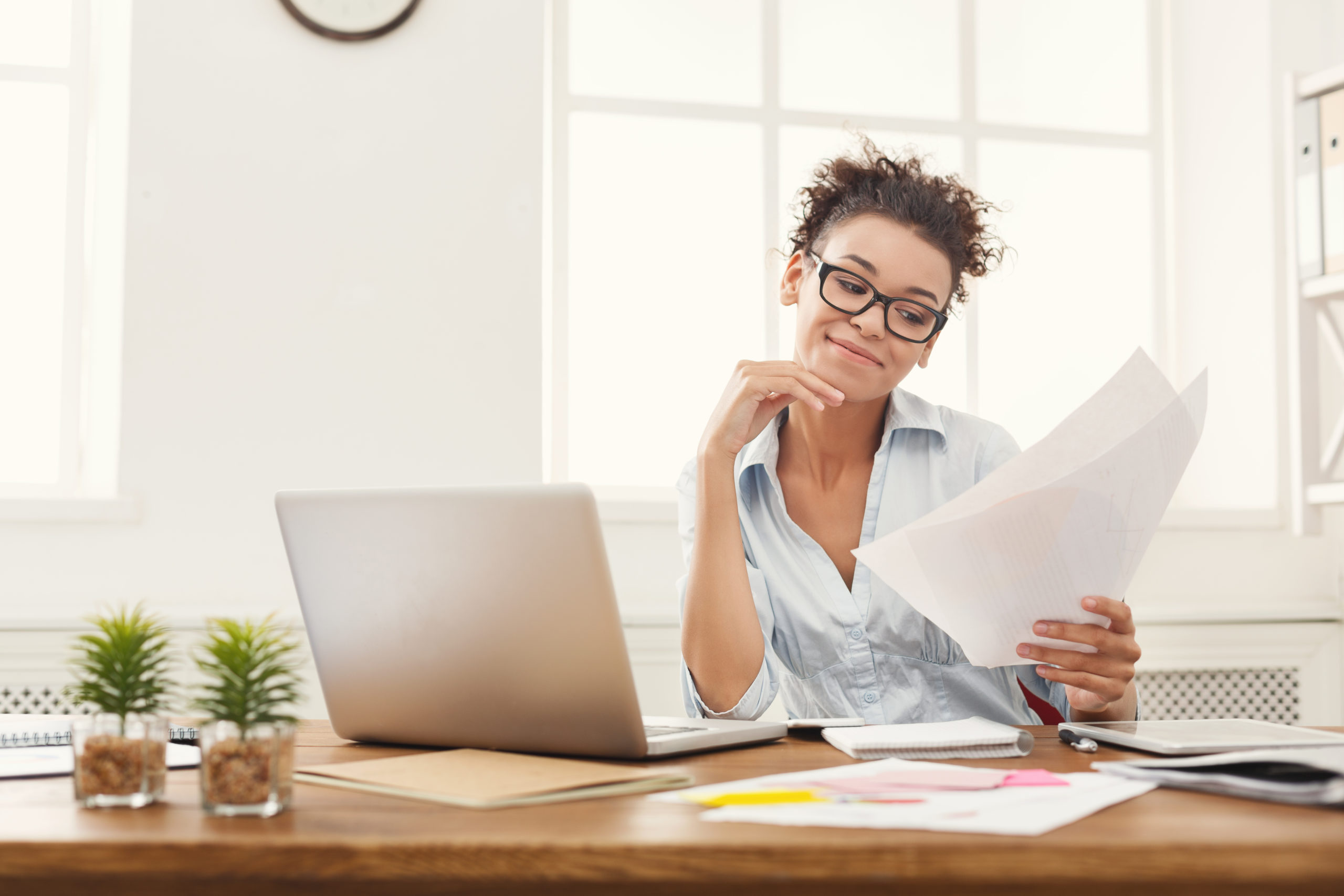 Young lady with slight grin holding and reviewing papers in front of laptop